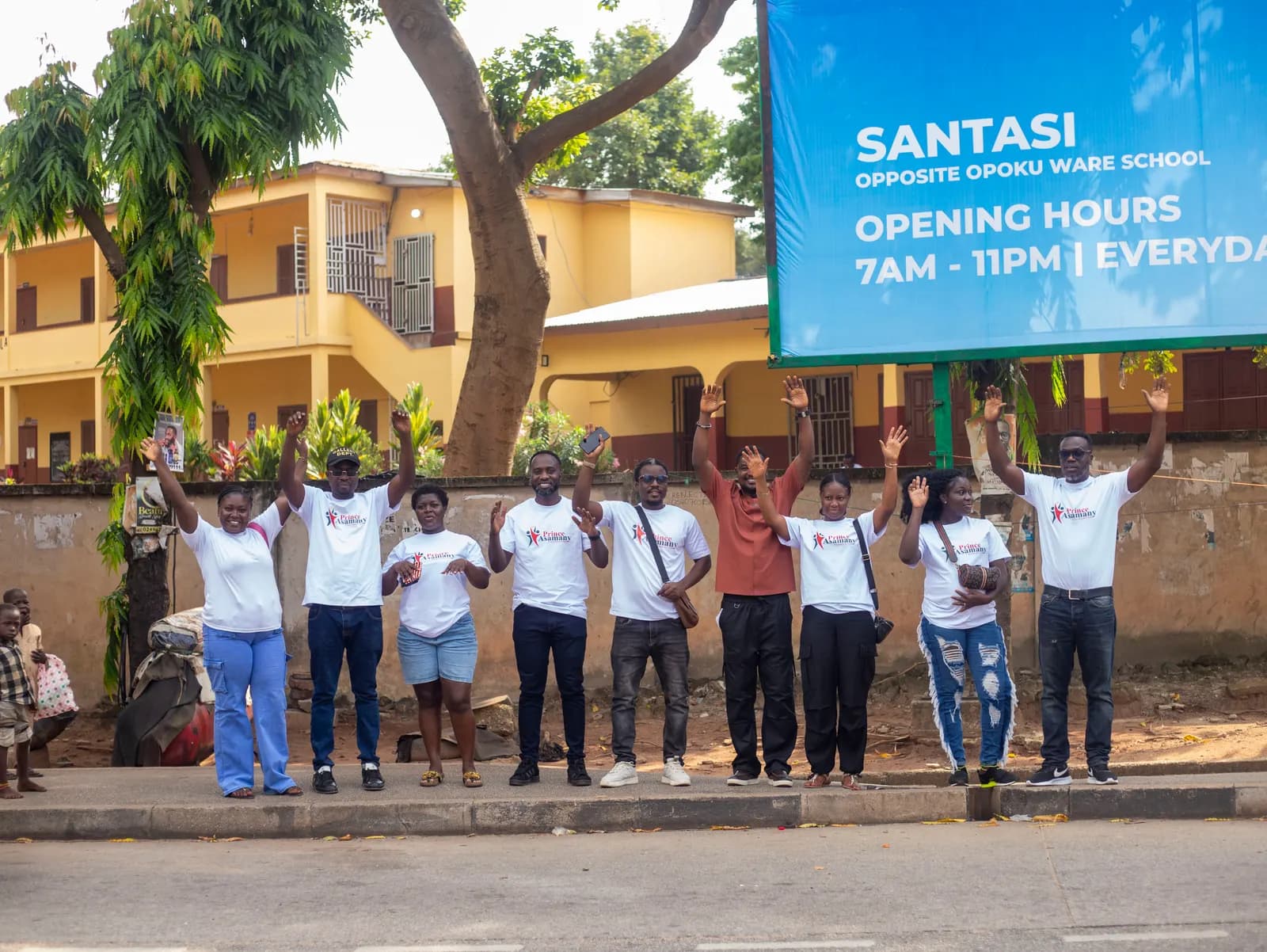 Foundation volunteers in Prince Asamany Foundation shirts celebrating with raised arms outside the Santasi event centre in Kumasi