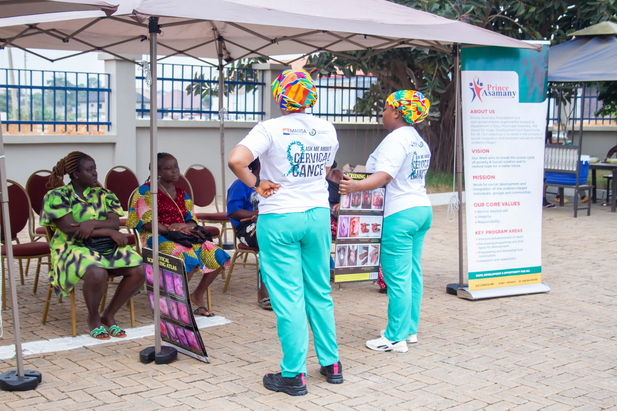 Women seated under a canopy awaiting free health screenings beside a Prince Asamany Foundation banner in Ejisu